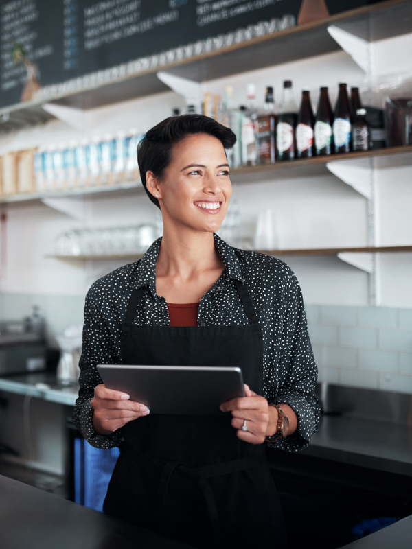 Eine Frau steht in einer Kaffeeküche mit Schürze und hält lachend ein Tablet in der Hand.
