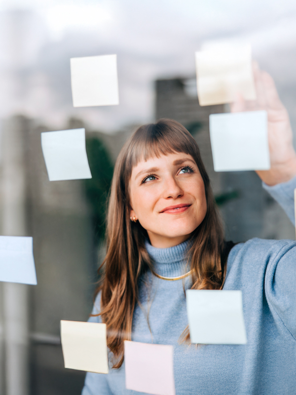 Eine junge Frau mit blondem Haar klebt lächelnd bunte Post-its an eine Fensterscheibe.